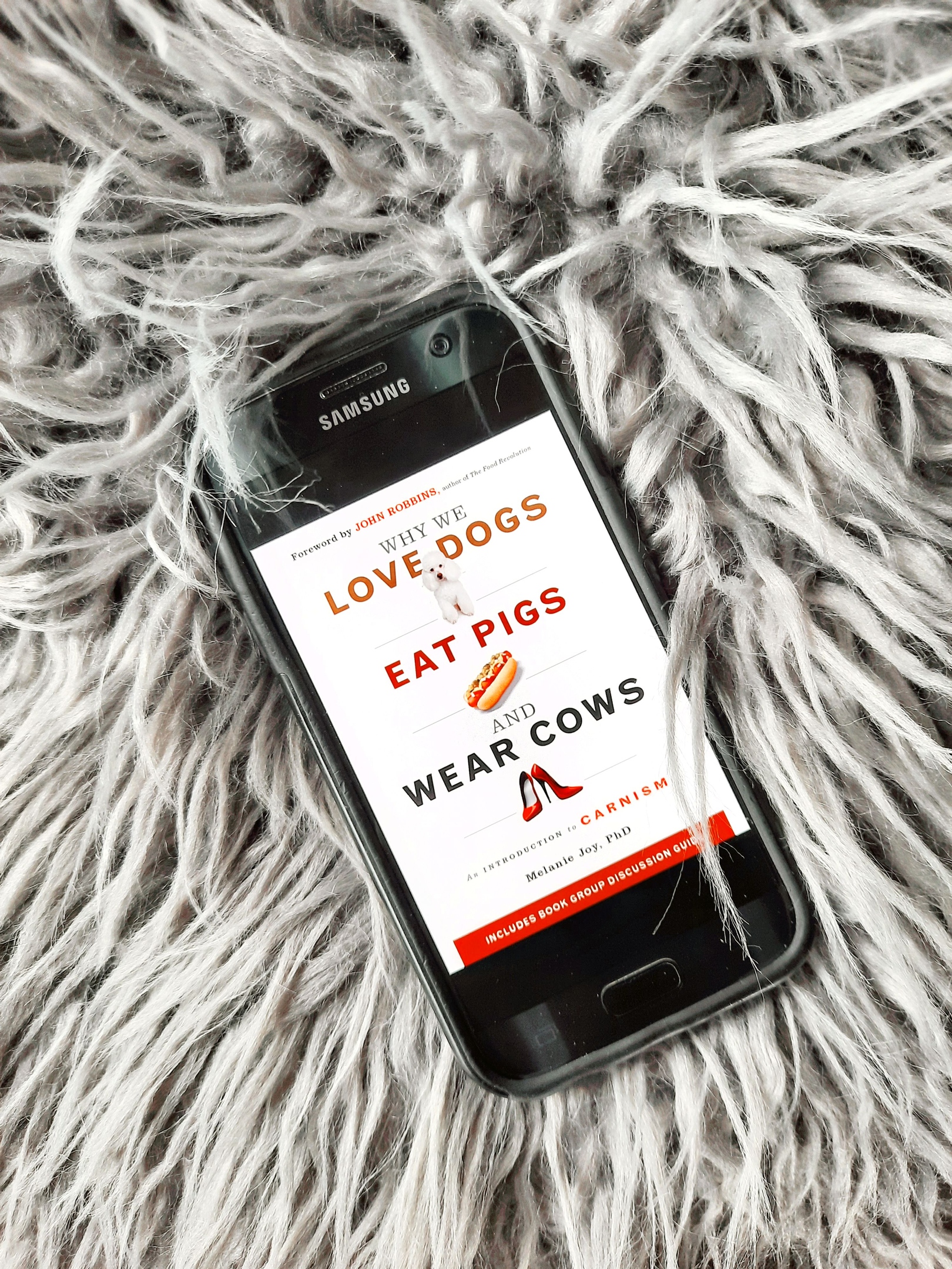 A phone lays atop a fluffy gray surface. The phone's screen shows the e-book cover of Why We Love Dogs, Eat Pigs, and Wear Cows.